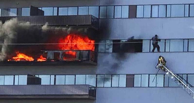 A man clinging to the exterior of the building on the sixth or seventh floor while a firefighter on a ladder attempts to rescue him—he was ultimately brought down safely.
