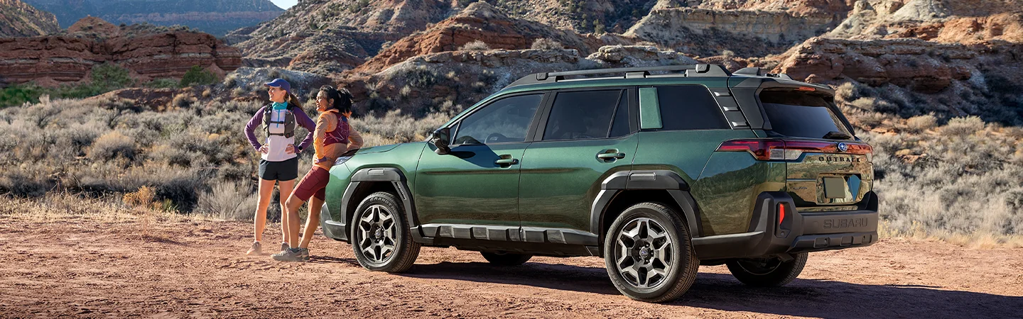 Side view of a parked 2026 Subaru Outback Wilderness in a desert with 2 wemon getting ready for a trail walk