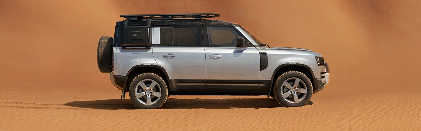 Side view of a parked silver 2025 Defender 110 in some sand