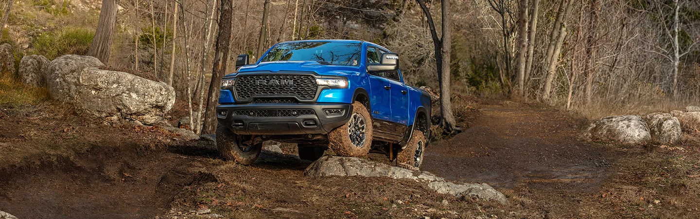 Front view of a blue 2026 RAM 1500 driving over some rocks in a forest