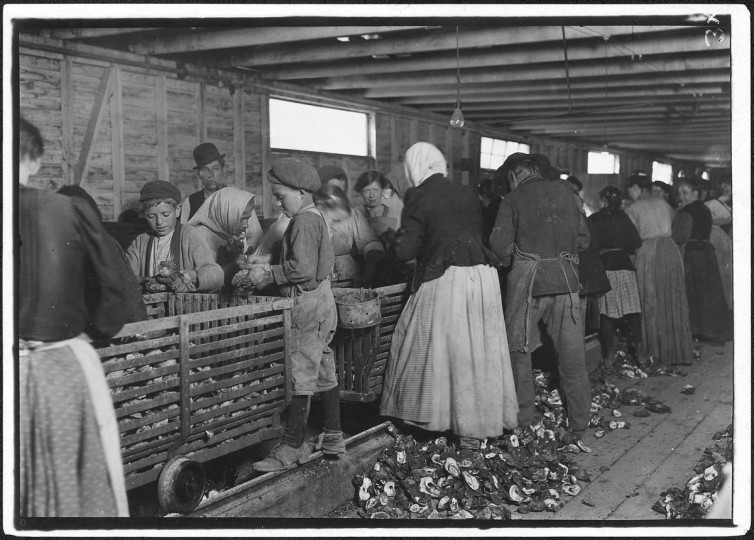 Original Caption: Johnnie, a nine year old oyster shucker. Man with pipe is a padrone who has brought these people from Baltimore for four years. He said, I tell you I have to lie to 'em. They're never satisfied. Hard work to get them. He is the boss of the shucking shed. Dunbar, La, March 1911. (Lewis Hine/Photo courtesy of NARA)