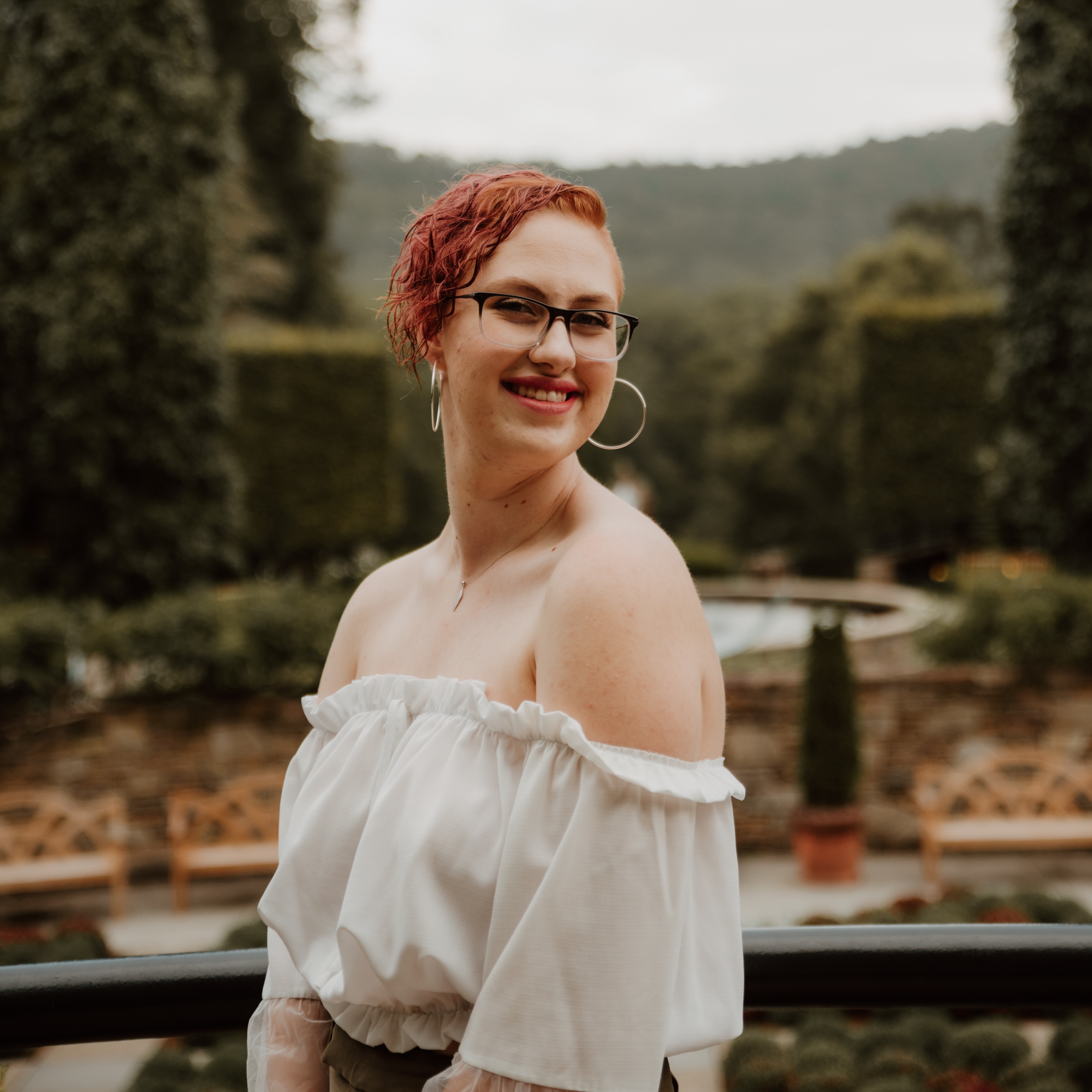 Person with red hair and glasses in a garden with topiary and hills in the background.