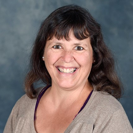 A woman with brown hair smiles warmly at the camera, wearing a beige cardigan over a dark top, set against a textured gray background.