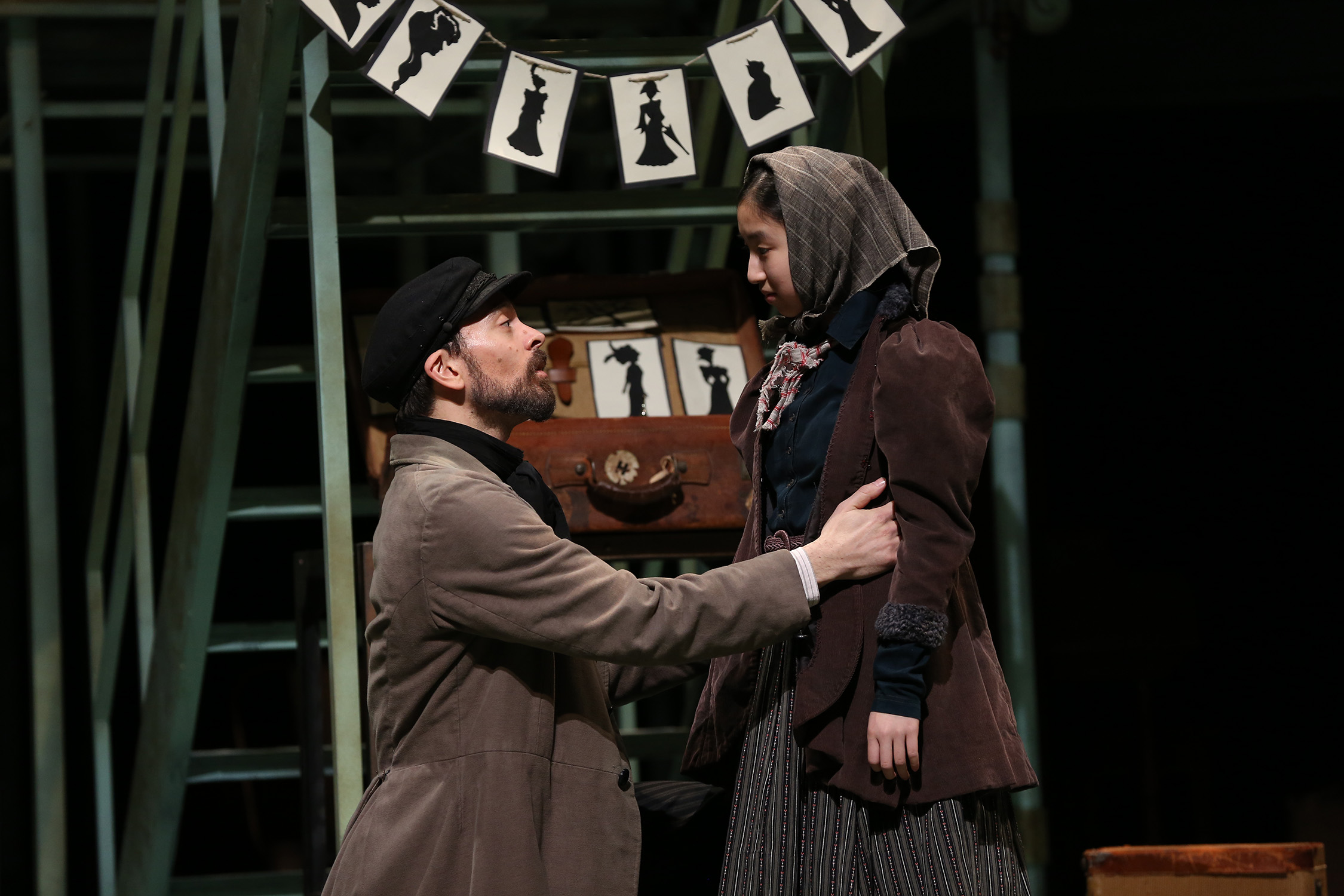 Photo of two actors: one man (Jonathan Atkinson) and one young girl (Dulcey Pham). They are dressed as Jewish immigrants in modest clothing. The young girl wears a headscarf. They are standing near a retail kiosk where the man sells silhouette portraits. Image is from the Ford's production of "Ragtime."