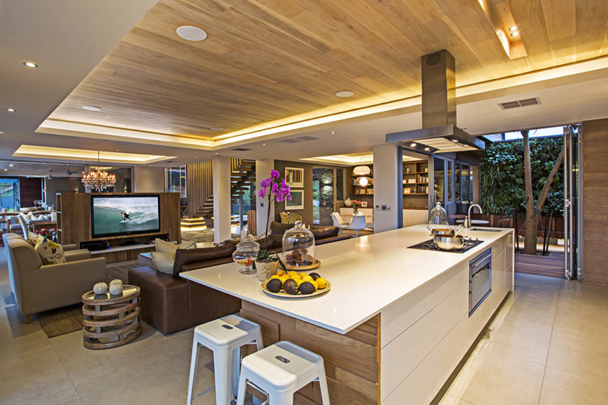 The kitchen sits within a vast open space area, defined by its large island and use of bright natural wood tones. The island features a built-in oven, range, sink, and space for dining.