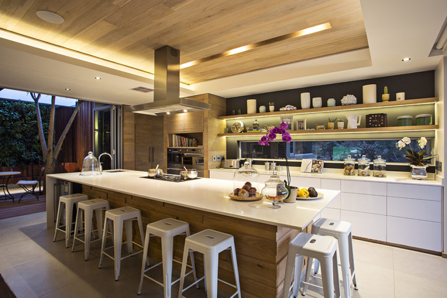 The kitchen also boasts sleek, hardware-less white cabinetry and intricate built-in shelving. The natural wood panels wrap the island, pantry to the left, and span the ceiling.