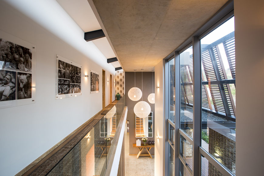 Moving up the stairs, we can see down into the main entryway and living room area from the catwalk with glass balustrade. Artwork spans the vast white walls on this floor, naturally lit via huge glass panels on the front.