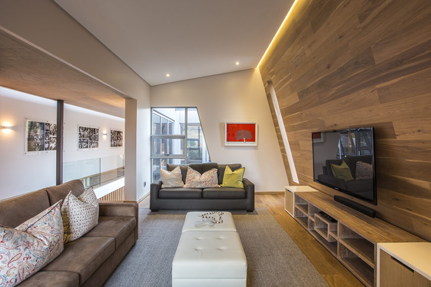 On the upper floor, we see an angular-shaped smaller living room area that flanks the entry space. Here, sleek wood paneling covers the wall and floors, while broad openings allow for views outdoors and throughout the interior of the home.