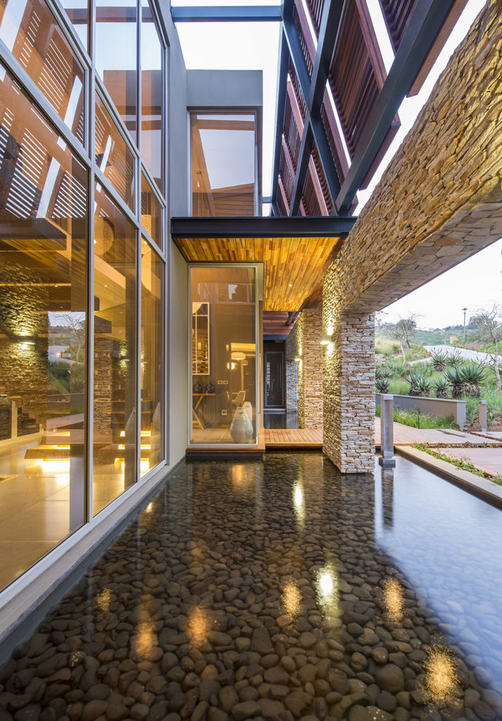 Just outside the main entry, we see a stone-bottom pond that flanks the wooden entry bridge, overlooked by massive glass panels on the home. Above, wooden louvered panels grant privacy and shade to the upper levels.