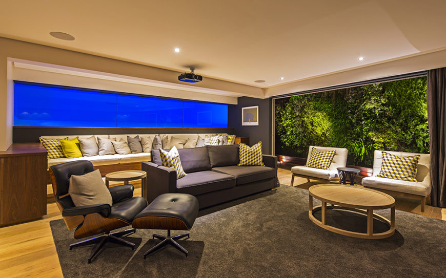 On this level we see another large living room space, with a lengthy sectional against the back wall and set of midcentury modern chairs in the foreground. To the right, another vast window offers views of the environment.