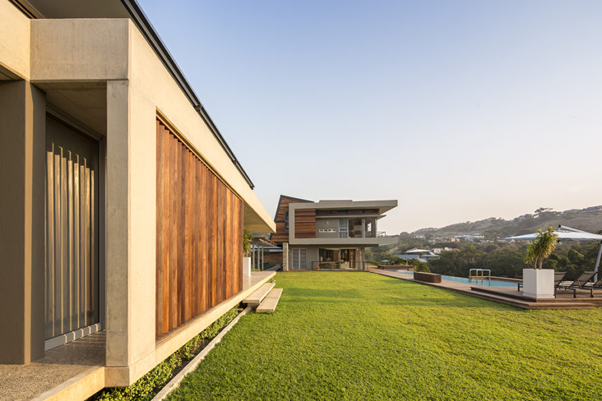 Moving to the expansive backyard, we see a minimalist landscape with lengthy pool to the right, wrapped in more sleek wood flooring. The building itself features numerous natural wood louvers for shade and privacy.