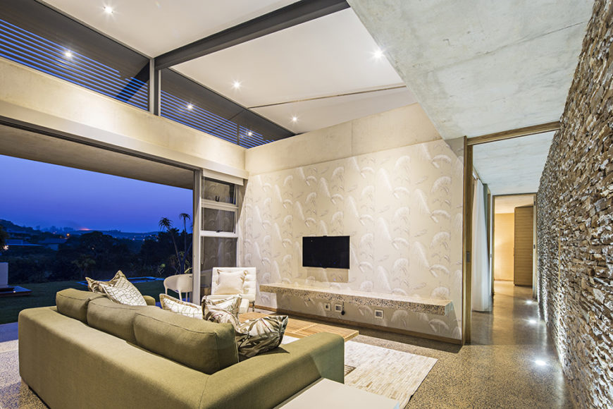 Within the numerous private and social spaces of the home, we see direct and broad access to the outdoors via sliding glass panels. Here, a stone wall connects all of the various rooms in the open plan design.
