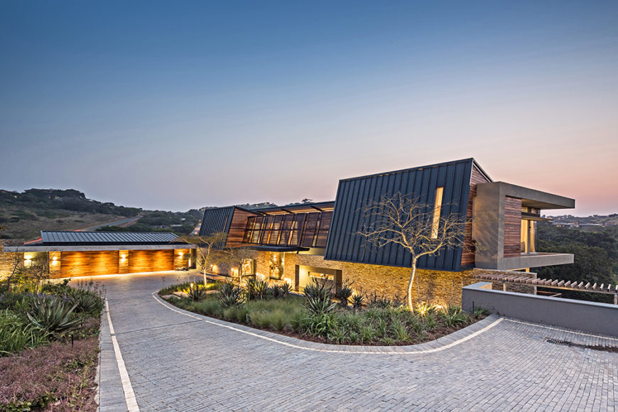 The brick driveway offers a sloped approach to the home, which glows at night via subtle exterior lights. From here we can see the vast landscape that the building overlooks.
