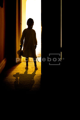 The silhouette of a little girl, in a night gown, holding a teddy bear, standing in a bedroom doorway.