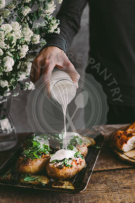 Man's hand pouring sour cream on baked stuffed potatoes with cheese, vegetables and rucola on wooden table. Rustic background