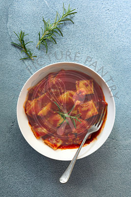 A bowl of ravioli in tomato and meat sauce with a fork on a blue textured background.