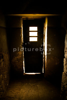 An old metal door, with a window, letting light into a damp and decaying room.