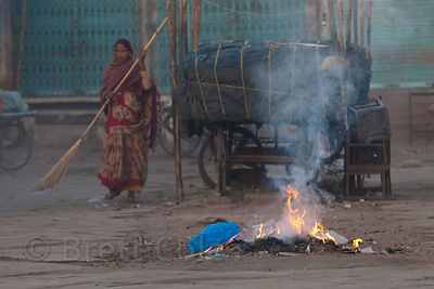 A woman sweeps the streets at sunrise admidst burning trash piles, Jodhpur, Rajasthan, India