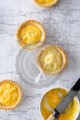 Three lemon tartlets with a bowl of lemon curd and a knife.
