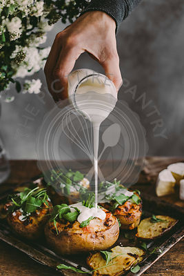Man's hand pouring sour cream on baked stuffed potatoes with cheese, vegetables and rucola on wooden table. Rustic background. Copy space