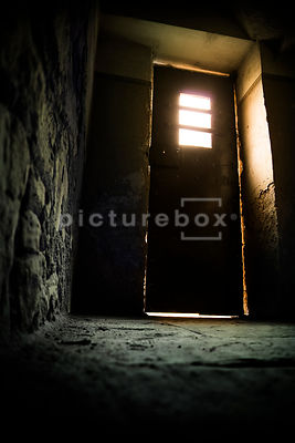 An old metal door, with a window, letting light into a damp and decaying room.