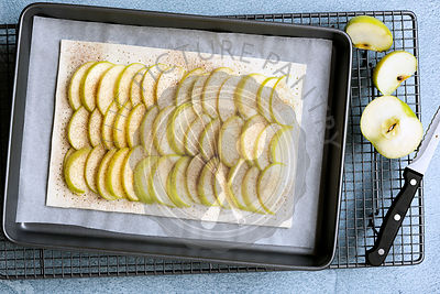 An uncooked rectangular tart with rows of apple on the pastry, a knife and cut pieces of cooking apple.