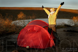 Backpacker waking up to sunny morning with arms raised by tent and lake