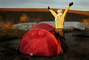 Backpacker waking up to sunny morning with arms raised by tent and lake