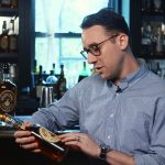 A young man wearing glasses sitting at a bar reading the label of a bottle of bourbon