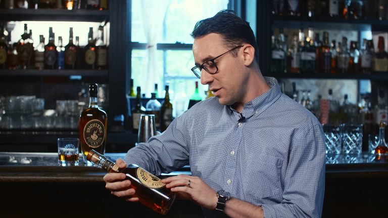 A young man wearing glasses sitting at a bar reading the label of a bottle of bourbon
