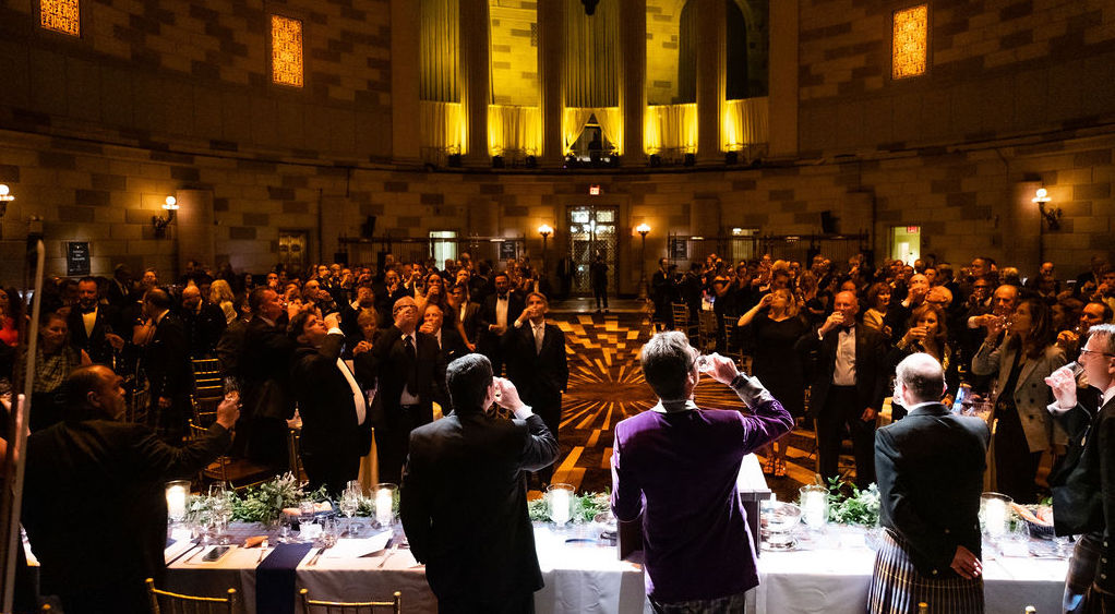A large oval room full of dinner guests with a head table drinking a toast