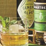 A cocktail in a rocks glass and garnished with a mint sprig sits in front of two bottles of chartreuse.