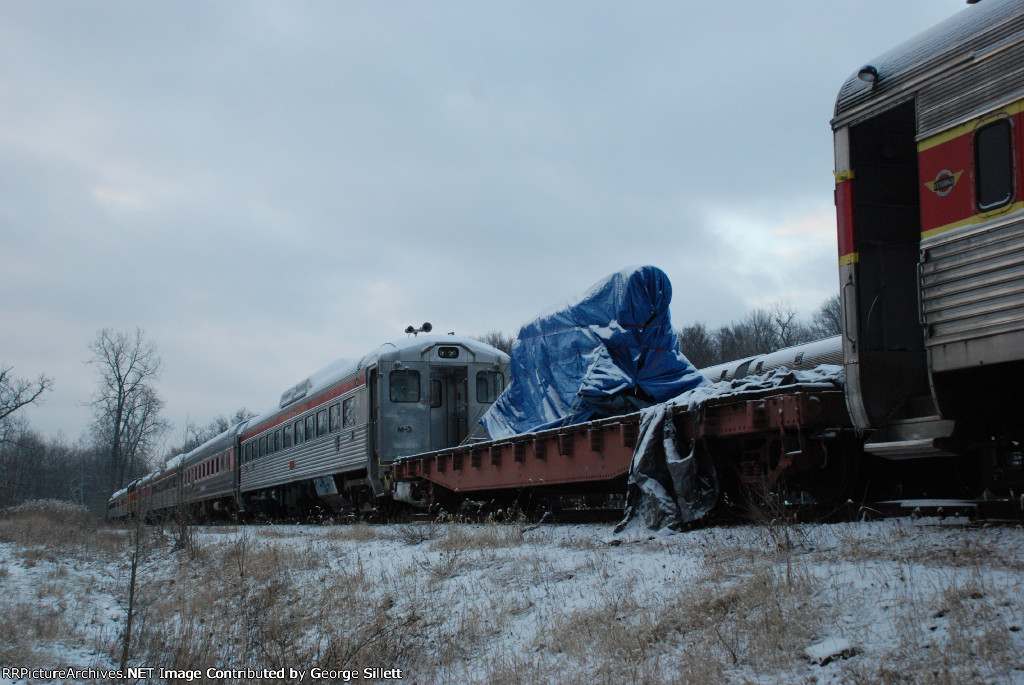 An engine on a flatcar, and several older RDC cars sit in teh storage line.