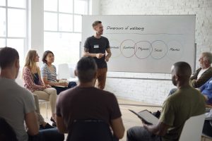A man stands in front of a whiteboard with Venn diagrams while speaking to a group of individuals seated in chairs.