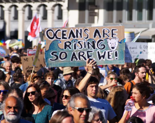 Climate protest with large crowd holding signs including "OCEANS ARE RISING SO ARE WE!" demonstrating environmental activism and concern about rising sea levels and climate change.