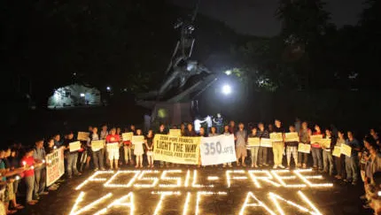 Nighttime climate protest with activists holding signs around illuminated "FOSSIL FREE VATICAN" text on ground, gathered near statue with bright lights creating dramatic scene advocating for environmental action.