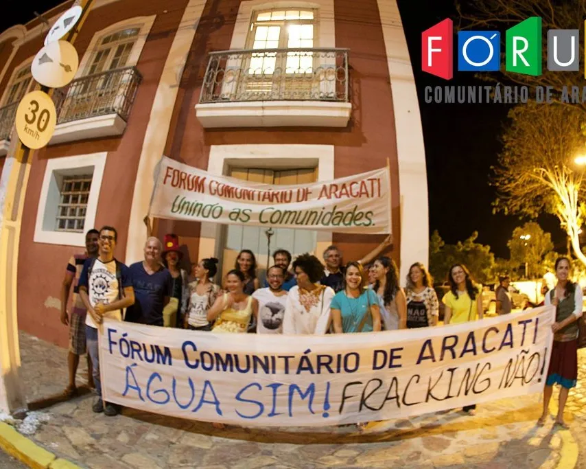 Community protest in Aracati, Brazil. Group holds banner reading "Fórum Comunitário de Aracati - Água Sim! Fracking Não!" (Water Yes! Fracking No!) in front of historic colonial building at night.