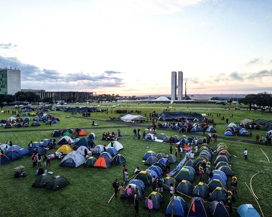 Hundreds of colorful tents pitched across a vast green lawn in front of Brazil's National Congress buildings in Brasília at sunset, with people gathering for what appears to be a large-scale protest or demonstration.