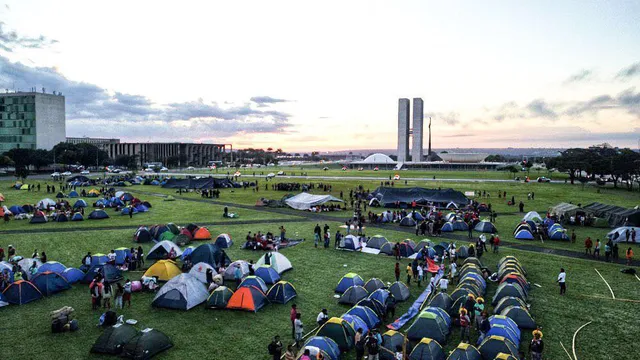 Hundreds of colorful tents pitched across a vast green lawn in front of Brazil's National Congress buildings in Brasília at sunset, with people gathering for what appears to be a large-scale protest or demonstration.