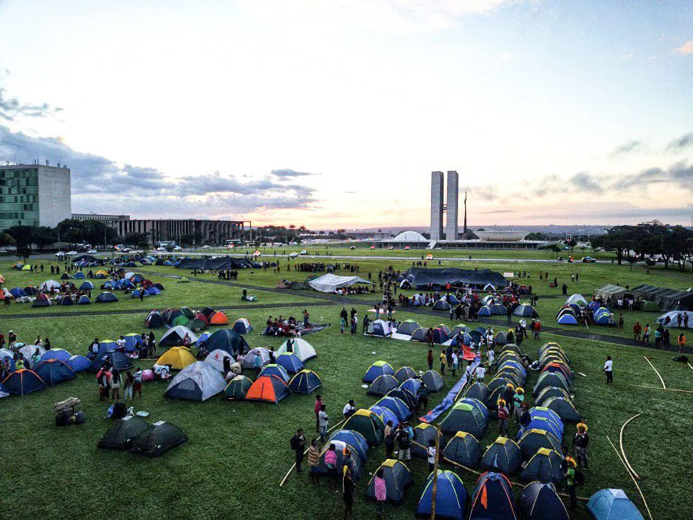 Hundreds of colorful tents pitched across a vast green lawn in front of Brazil's National Congress buildings in Brasília at sunset, with people gathering for what appears to be a large-scale protest or demonstration.