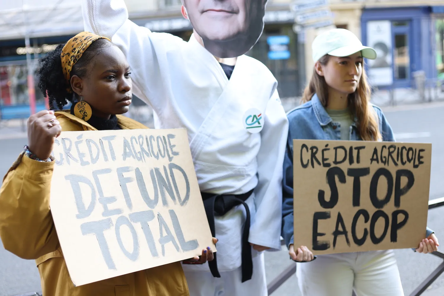 Two protesters demonstrate against Crédit Agricole bank, holding signs demanding "DEFUND TOTAL" and "STOP EACOP" in front of a large advertisement featuring a bank employee in white uniform.