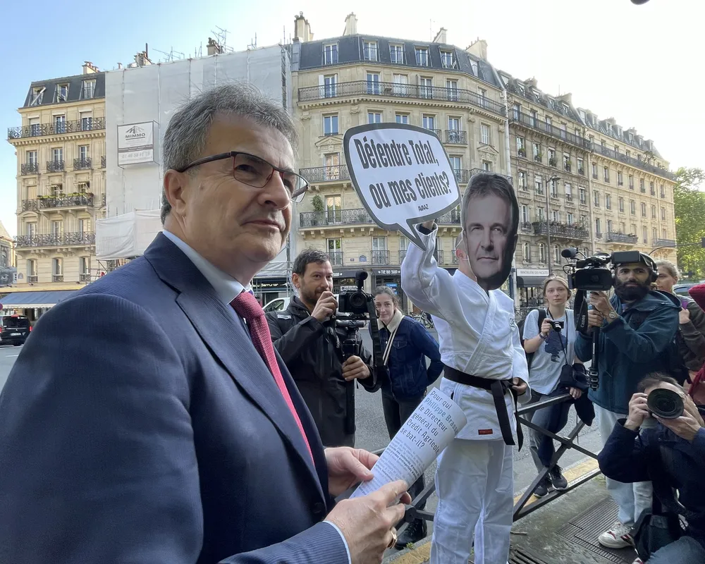 A man in a suit stands before media cameras while a protester behind him wears a mask and holds a sign in French on a Paris street with classic Haussmann buildings in the background.