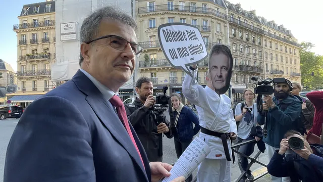 A man in a suit stands before media cameras while a protester behind him wears a mask and holds a sign in French on a Paris street with classic Haussmann buildings in the background.