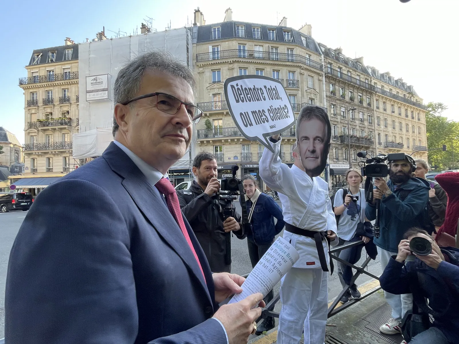 A man in a suit stands before media cameras while a protester behind him wears a mask and holds a sign in French on a Paris street with classic Haussmann buildings in the background.
