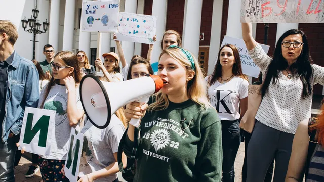 Young woman in green sweater holding megaphone and speaking to crowd outside