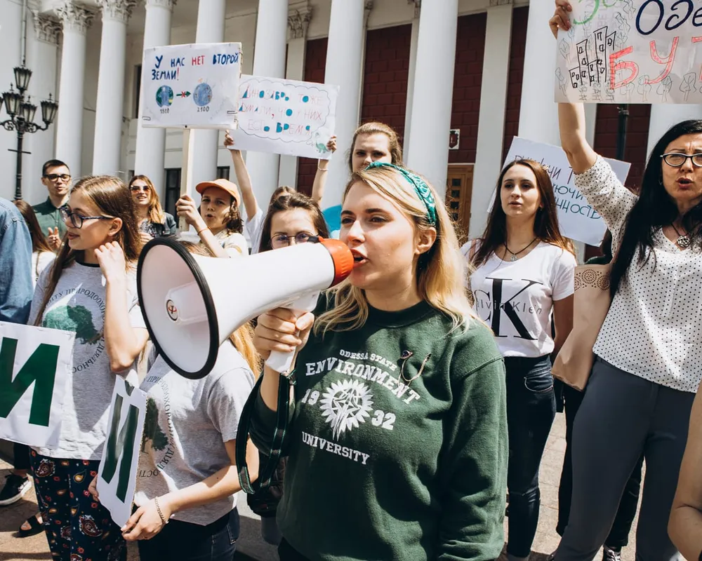 Young woman in green sweater holding megaphone and speaking to crowd outside