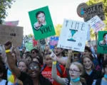 Protesters gather at a demonstration holding signs, including one with a green background. The crowd appears diverse with people of various ages participating in what seems to be a public rally or march.