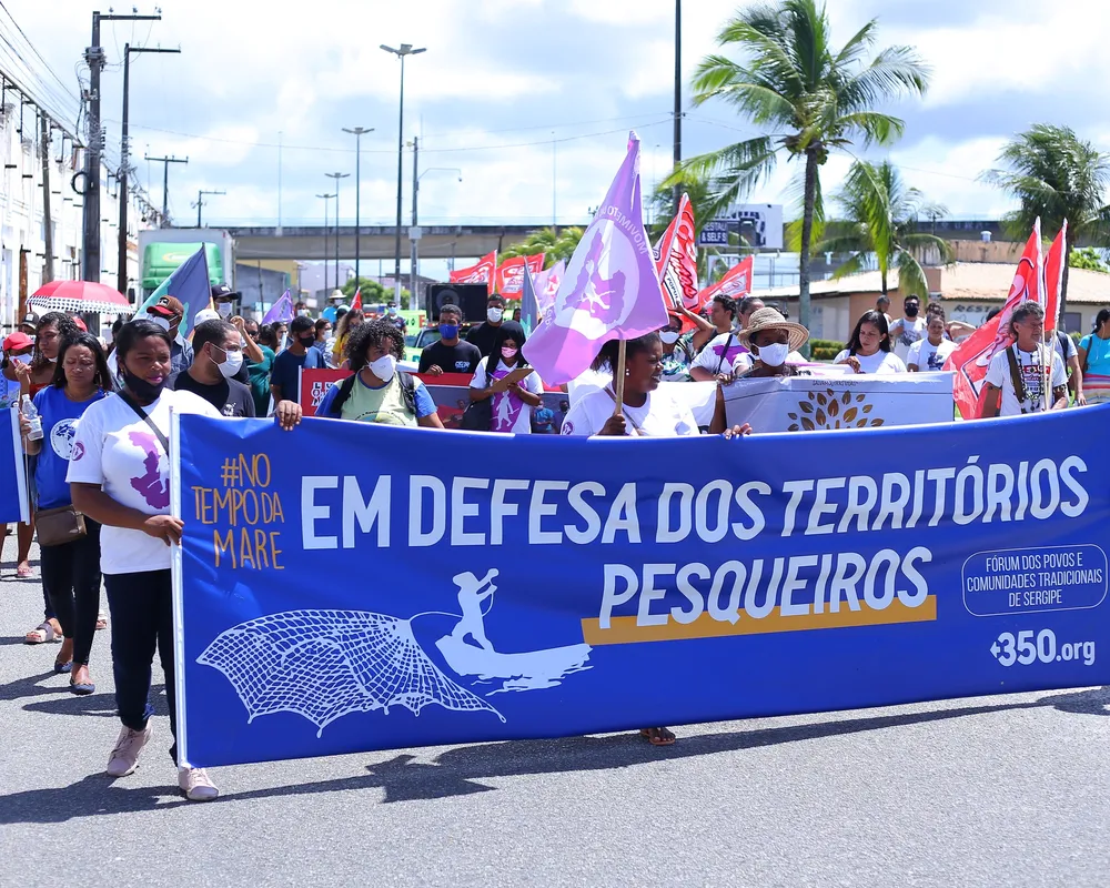 A protest march with people carrying a large blue banner reading "EM DEFESA DOS TERRITÓRIOS PESQUEIROS" (In Defense of Fishing Territories). Palm trees and urban infrastructure visible in background under cloudy skies.