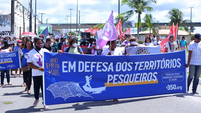 A protest march with people carrying a large blue banner reading "EM DEFESA DOS TERRITÓRIOS PESQUEIROS" (In Defense of Fishing Territories). Palm trees and urban infrastructure visible in background under cloudy skies.