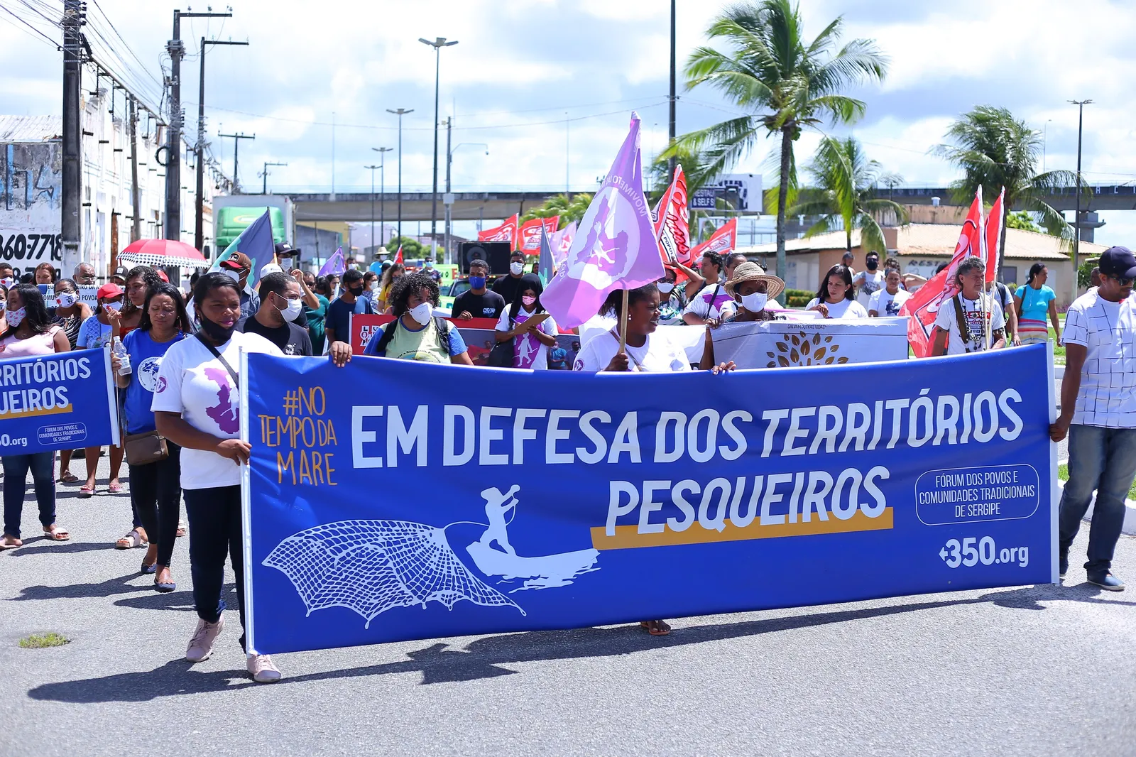 A protest march with people carrying a large blue banner reading "EM DEFESA DOS TERRITÓRIOS PESQUEIROS" (In Defense of Fishing Territories). Palm trees and urban infrastructure visible in background under cloudy skies.